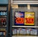 An advertisement display in a store window features lottery jackpot amounts for Mega Millions and Powerball, both showing jackpots of 208 million and 200 million respectively. Below the display, several lottery tickets and promotional posters are visible. To the left of the display, there is a refrigerator stocked with various drinks including different flavors of sports drinks and bottled water.