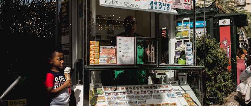 A child holding an ice cream cone stands outside a small kiosk or lottery ticket booth. The booth displays various colorful lottery tickets in the window. There are signs advertising a significant lottery prize amount in Spanish. A person can be seen partially obscured inside the booth, and plants are visible around the area, suggesting an outdoor setting.