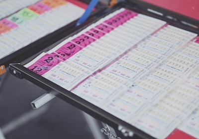 A collection of lottery tickets is organized neatly in a metal tray. The tickets display various numbers and have color-coded sections, primarily in pink and white. Some tickets have vibrant green sections, adding contrast to the overall layout.