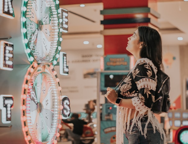 A woman is standing in an arcade looking at a large colorful game machine with bright lights. The game machine features large spinning wheels labeled with 'tickets' and has a vibrant design. The woman is casually dressed in a jacket with fringe details and is holding something in her hand while wearing a smartwatch.