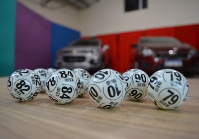 A collection of white lottery balls with black numerals scattered on a wooden surface. Two vehicles are blurred in the background, parked in a room with multicolored walls including teal, purple, blue, and red panels.